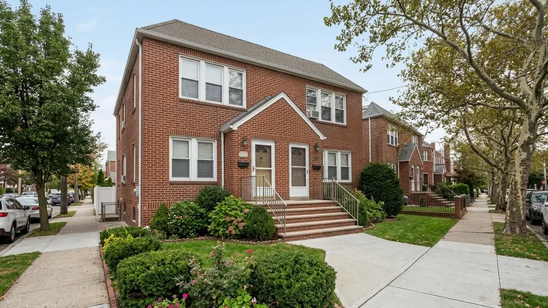 A brick two-family home in Queens with a small front yard, a stoop, and a swept front walk in daylight